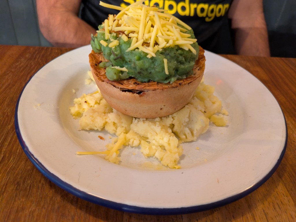 A pie on top of mashed potatoes, topped with mushy peas and grated cheddar