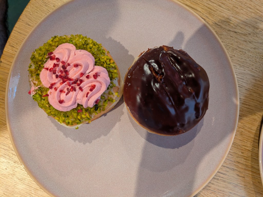Two filled donuts on a plate. One is topped with green icing and chopped pistachios and pink raspberry cream. The other is topped with chocolate.