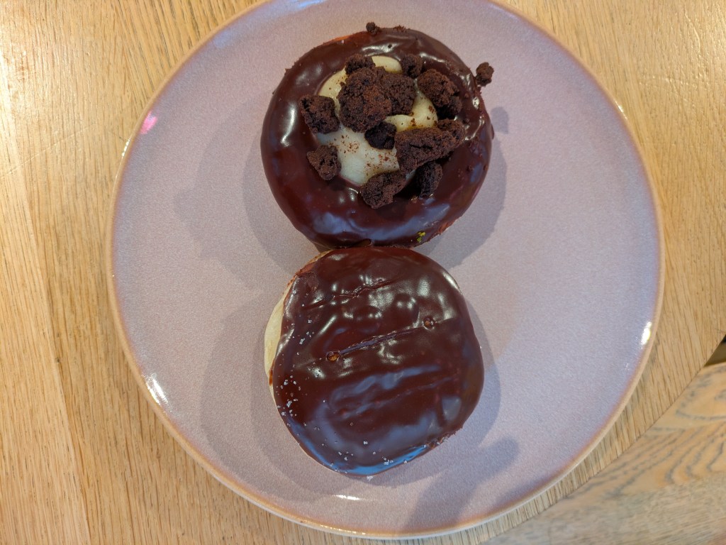 Two donuts on a plate, one a ring glazed with chocolate and topped with a blob of cream cheese ice cream and some biscuit crumbs, the other a filled donut topped with chocolate.