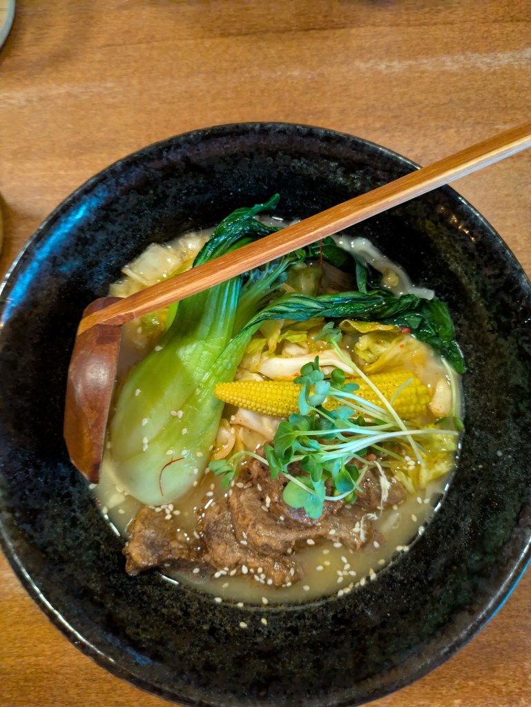 A bowl of ramen with a creamy broth, some big thick slices of vegan beef, a piece of baby sweetcorn and a large pak choi