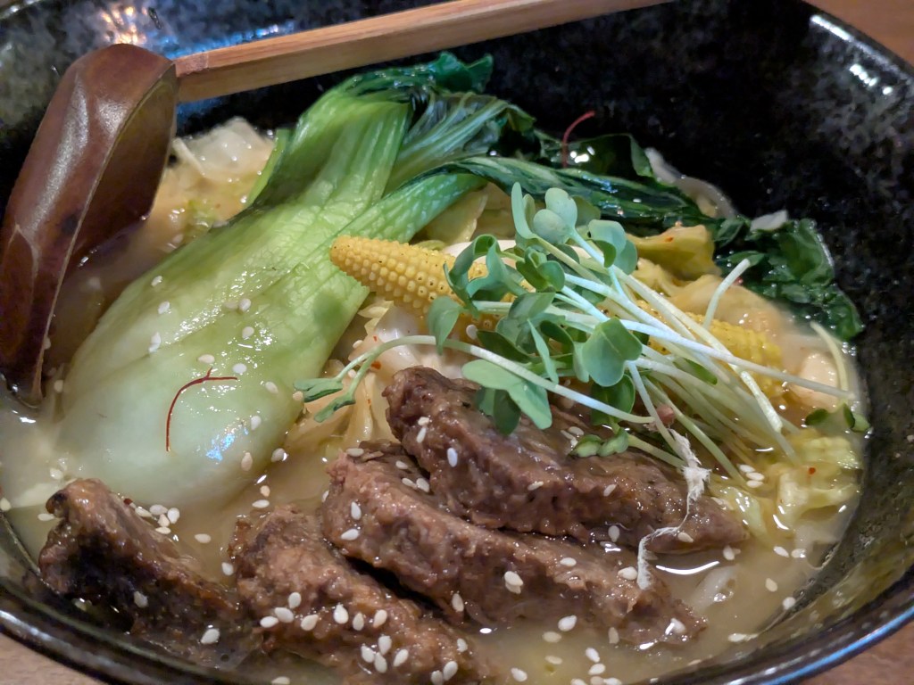 A bowl of ramen with a creamy broth, some big thick slices of vegan beef, a piece of baby sweetcorn and a large pak choi
