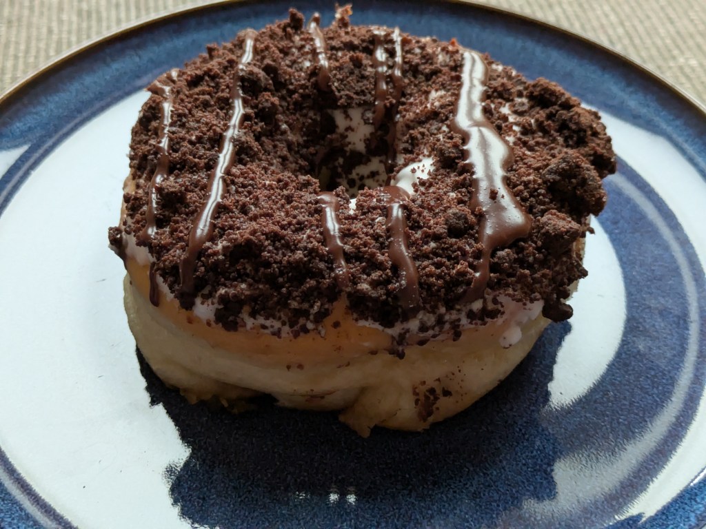A ring donut with white icing barely visible under a chocolate biscuit crumb coating.