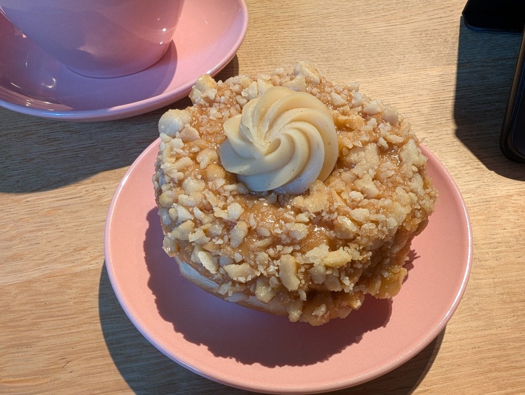 A ring donut topped with a sticky looking caramel glaze and chopped nuts, with a blob of cream cheese icing in the middle