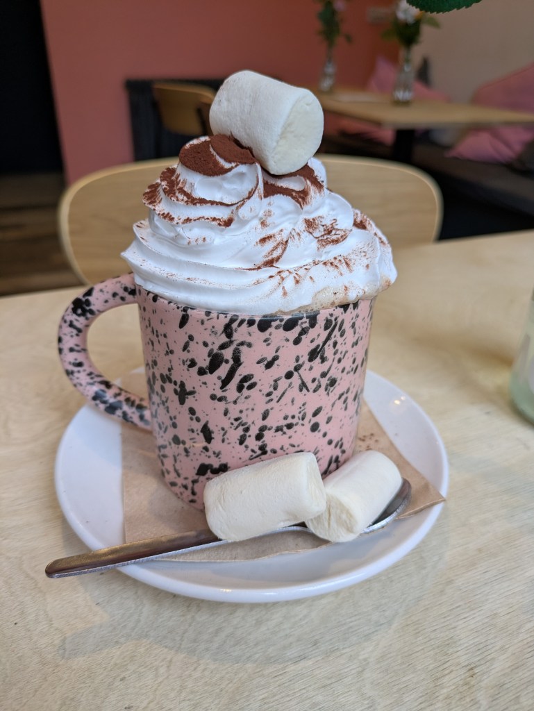 A mug of hot chocolate topped with a large swirl of whipped cream on top and several large marshmallows in and around the mug.