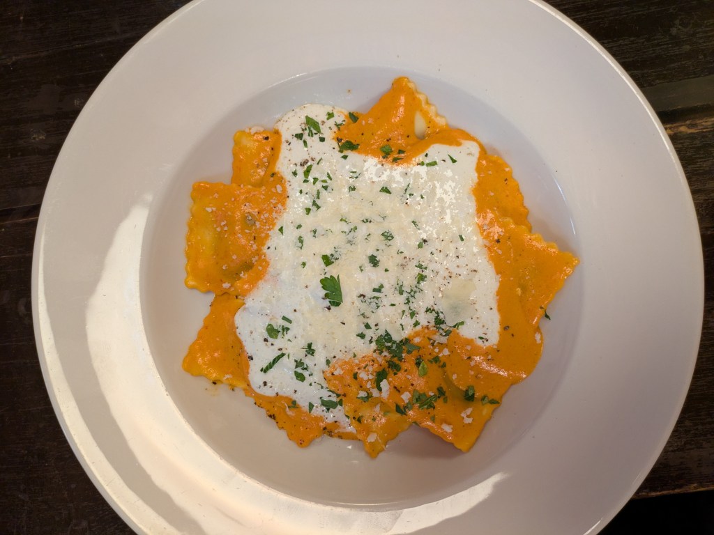 A dish of ravioli in an orange sauce with white cheese sauce on top