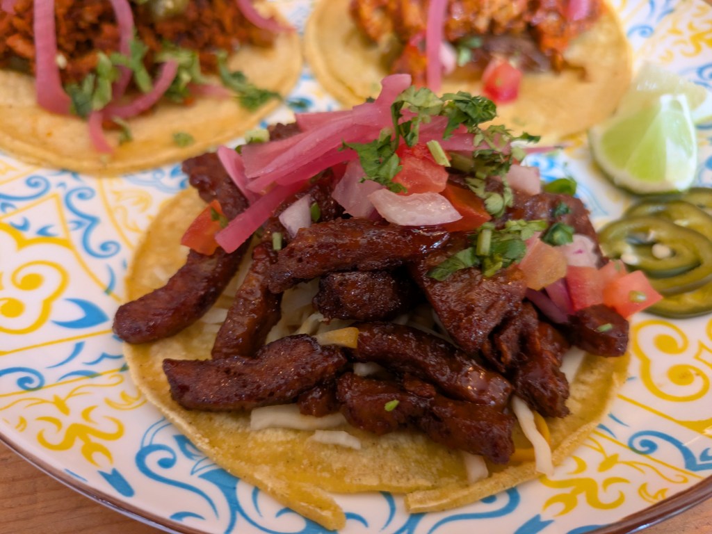 A taco filled with crispy strips of meat with a sticky glaze, garnished with some pickled and fresh vegetables