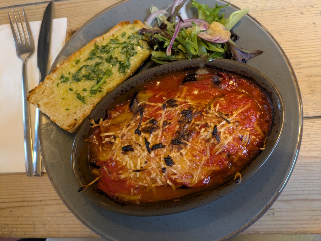 A dish of aubergine parm topped with melted cheese, served alongside a small side salad and a big slice of buttery garlic bread