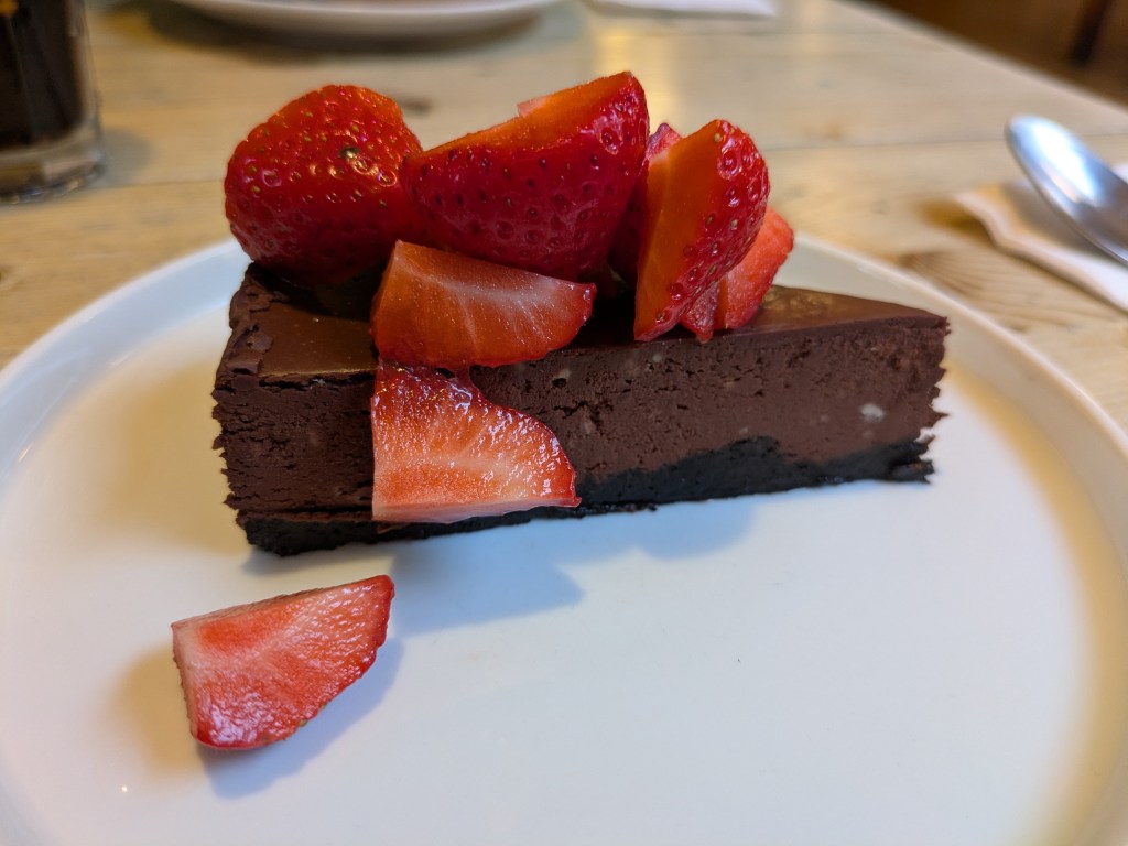 A slice of chocolate cake with a hard base topped with chocolate ganache, garnished with strawberries