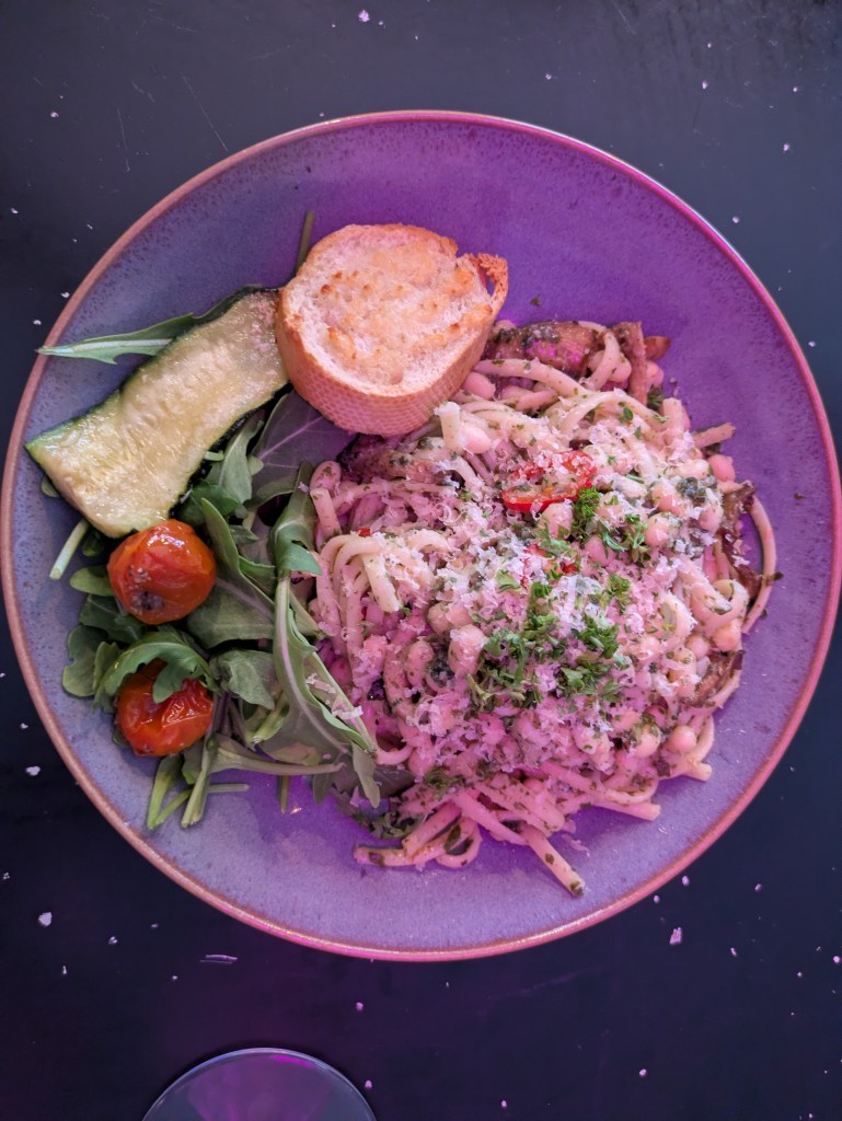 A bowl of linguini with a creamy sauce and various toppings, alongside a piece of garlic bread and a side salad