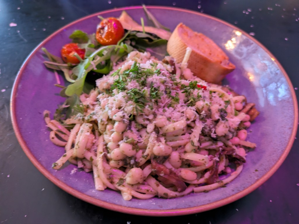 A bowl of linguini with a creamy sauce and various toppings, alongside a piece of garlic bread and a side salad
