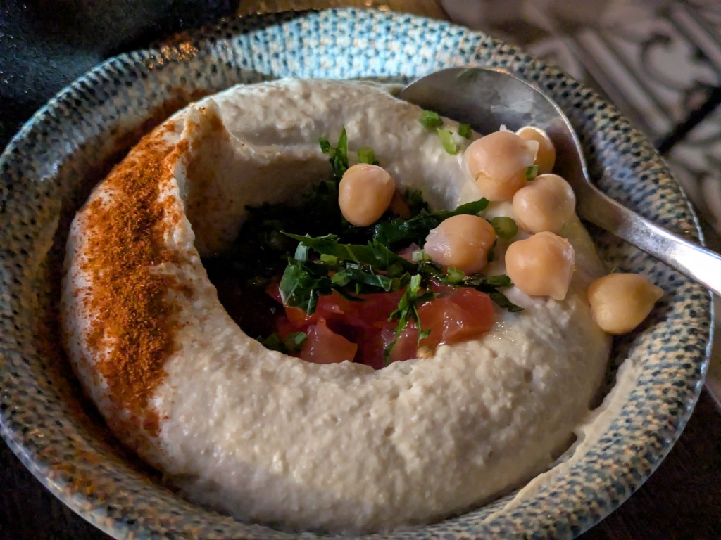 A dish of hummus, garnished with chickpeas, veg, and spice