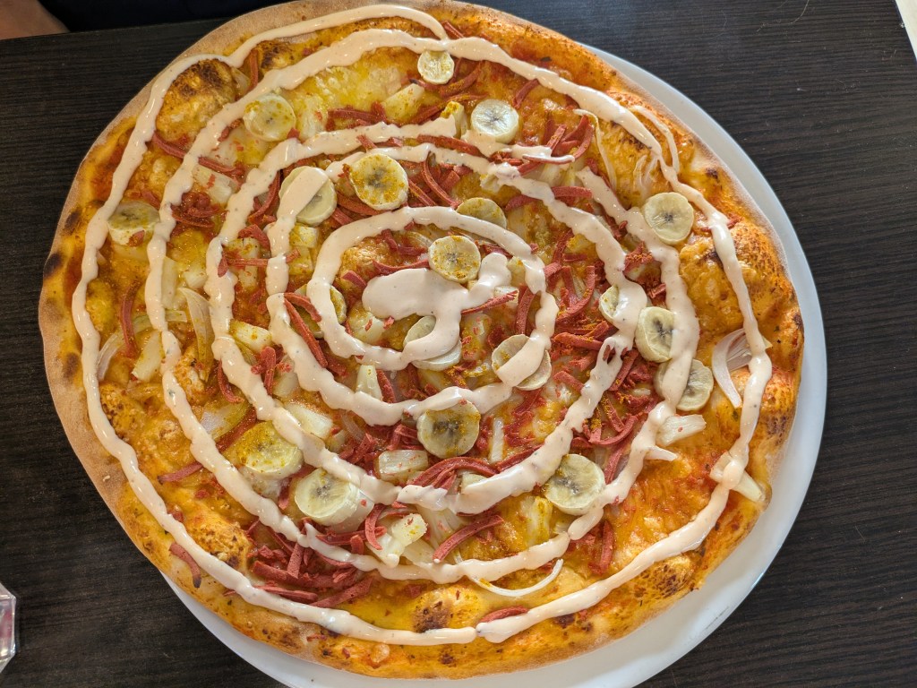 A pizza with lots of toppings including round banana slices, covered with a spiral of sauce